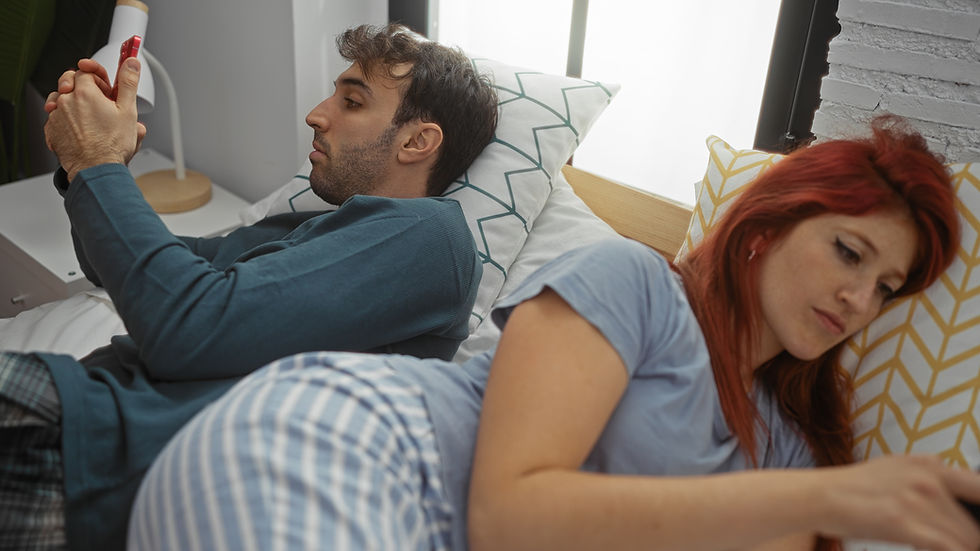 Couple lying back to back in bed, man and woman using phones, showing distance in their relationship, with modern bedroom setting suggesting indoor lifestyle.

