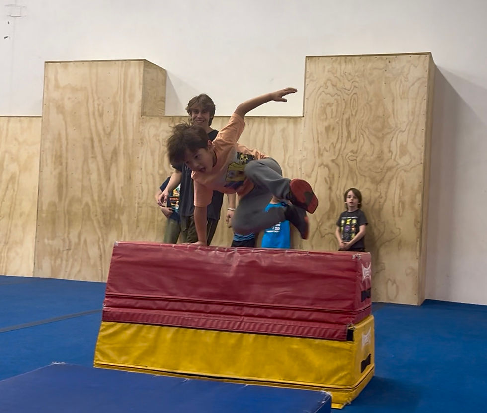 A boy in an orange shirt jumps over a red and yellow mat in a gym with wooden walls, while others watch, conveying excitement and energy.