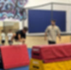 A boy in black vaults over red foam blocks in a gym. A coach watches, impressed.