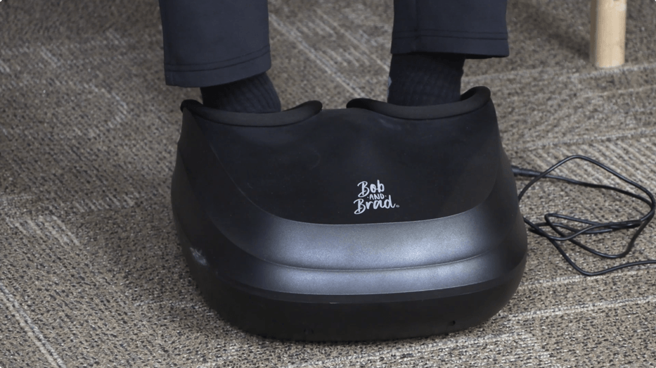 A black Bob and Brad Foot Massager is sitting on the floor, with a person's feet inside.