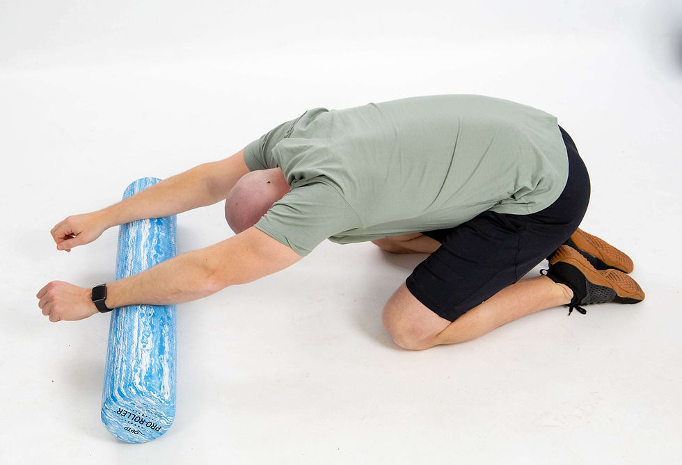 Man kneeling on the floor in a child's pose stretch, sitting back toward his heels with both arms extended forward over a blue foam roller and his head lowered toward the floor to stretch his shoulders and upper back.