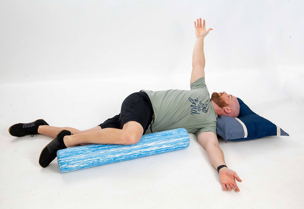 Man lying on his back with his head supported by a pillow, one knee resting across a blue foam roller and one arm extended upward while the other rests on the floor, performing a thoracic spine rotation stretch.