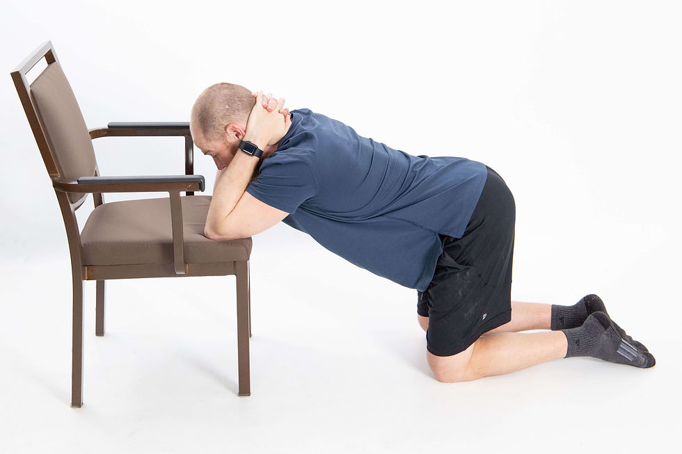 Man kneeling on the floor with his elbows resting on a chair seat, hands clasped behind his neck, and chest lowered toward the floor to stretch his shoulders, upper back, and triceps.