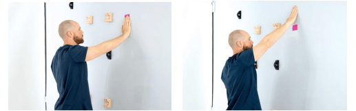 Mike is standing facing a wall. His right hand is on a piece of tape on the wall and then above the tape. The tape is just above eye level.