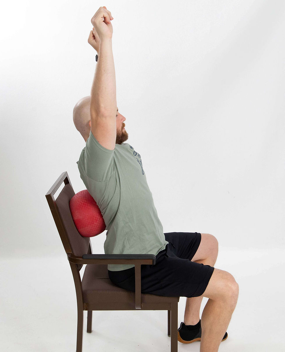 Side view of Mike seated in a chair with a red ball positioned between his upper back and the chair, leaning backward with both arms extended overhead to stretch his upper back.