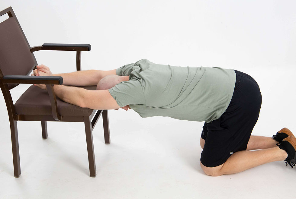 Man kneeling on the floor with his hips pushed back, arms extended forward onto a chair seat, and head lowered between his arms to stretch his shoulders and upper back.