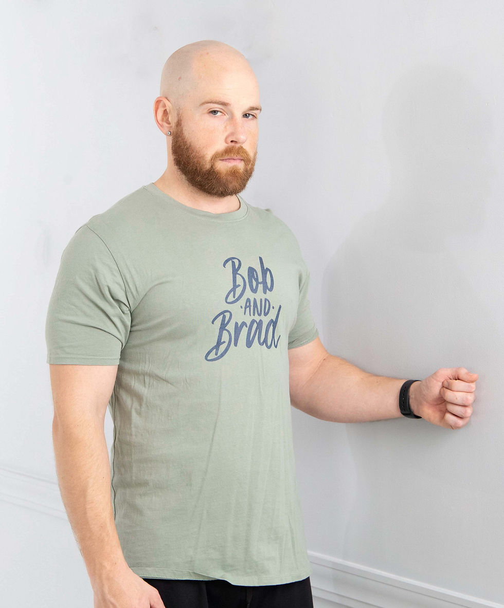 Mike is wearing a sage green Bob and Brad T-shirt, standing beside a wall, pressing his wrist against the wall with his elbow bent at approximately 90 degrees to perform a shoulder isometric exercise.
