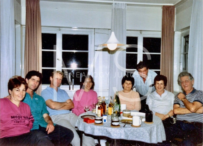 Eight people gather around a table with drinks and snacks in a cozy room. They appear relaxed and cheerful, with curtains and a hanging lamp.