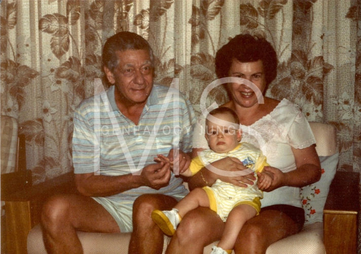 Elderly man and woman sit on a couch holding a baby in yellow. Floral curtains in the background. Warm, nostalgic atmosphere.