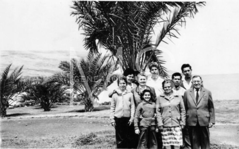 A group of nine people stand outdoors near palm trees. They are smiling, dressed in casual clothing. The background shows a landscape. Black and white.