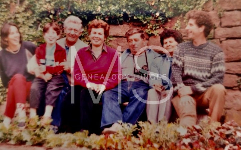 A group of seven people sit smiling on a stone bench outdoors, with ivy-covered walls in the background and text overlaying the image.