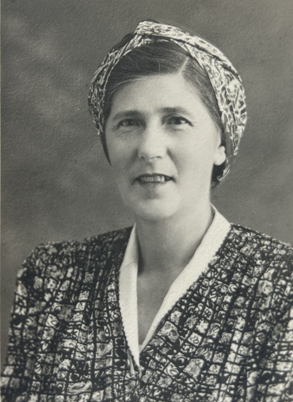 Woman with a patterned headscarf and dress, smiling slightly against a plain background. Black and white photograph.