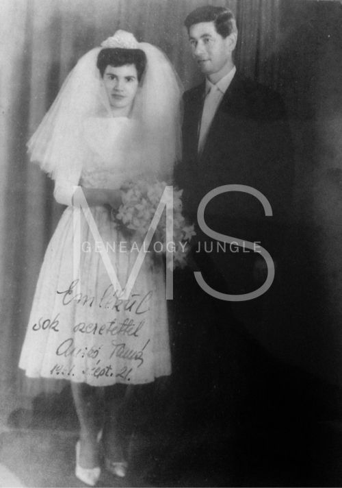 Bride in white dress and veil with groom in suit, holding flowers, pose in front of curtain. Handwritten note on dress, black and white photo.