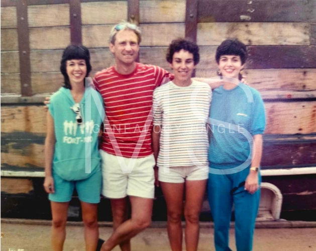 Four people smiling in casual clothes stand against a wooden wall. One wears a red striped shirt, others in blue and white outfits. Relaxed mood.