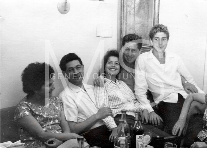 A group of five people sit closely on a couch, smiling. Bottles and cups are on the table. The setting is a cozy, indoor room.
