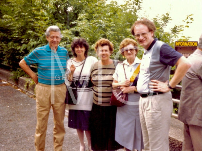 Five people smiling outdoors with greenery, wearing casual clothes. A yellow "INFORMATION" sign is visible in the background.