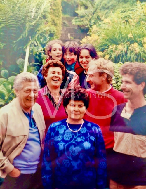 Smiling group of seven people outdoors with lush greenery. Bright clothing, joyful expressions.