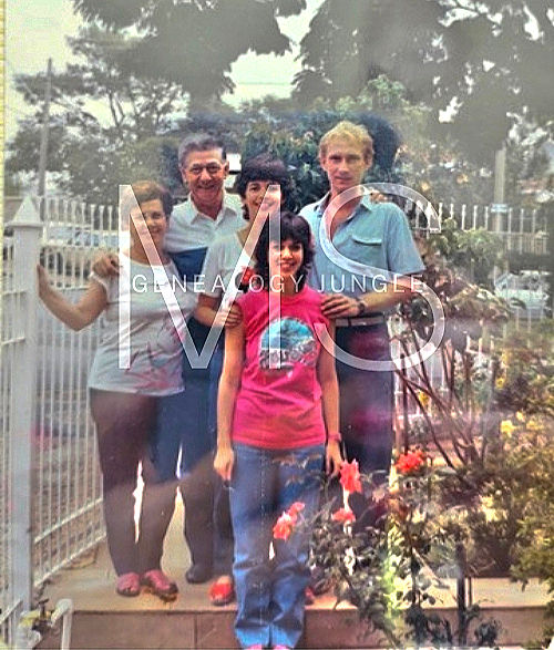 Five people smile in a garden, with trees and a white fence in the background. One wears a bright pink shirt.