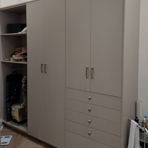 Large, beige-colored wardrobe with multiple compartments and drawers. The top section has two large doors with handles, the middle section has smaller compartments with various items inside, and the bottom section has several drawers with round knobs. On top of the wardrobe, there is a basket containing various bottles and a roll of paper towels. The floor is wooden with a herringbone pattern.