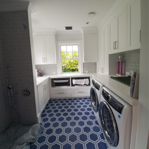 Shaker style laundry room with white cabinetry and silver handles, blue hexagonal tile floor, two front-loading washing machines or dryers on the right with a countertop above, window with greenery view, light gray subway tile walls, recessed lighting, storage baskets, a small sink area, and a dog bed in the corner.