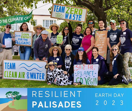 A large group of Resilient Palisades volunteers pose together outdoors under a leafy tree during the Earth Day 2023 celebration. They hold handmade signs for different teams, including Zero Waste, Clean Air & Water, and Solar Clean Energy for the Palisades. Several people wear navy Resilient Palisades T-shirts, some wear hats and sunglasses, and one person is dressed in a cow costume holding a “Save the Planet, Go Vegan” sign. A banner reading “Resilient Palisades Earth Day 2023” runs across the bottom of the image.