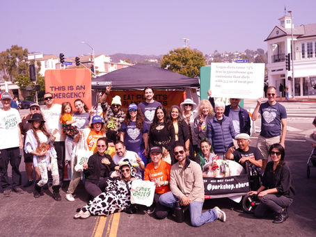 A large group of Resilient Palisades volunteers standing together outdoors on a street, holding signs and smiling at the camera during an Earth Day gathering. Buildings and a blue sky are visible in the background.