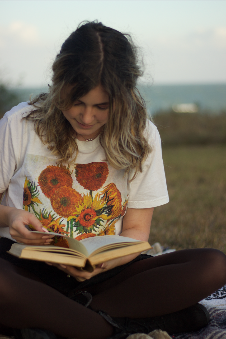 A photo of author Halli Castro sitting in a field reading a book, in a t-shirt with painted sunflowers on the front