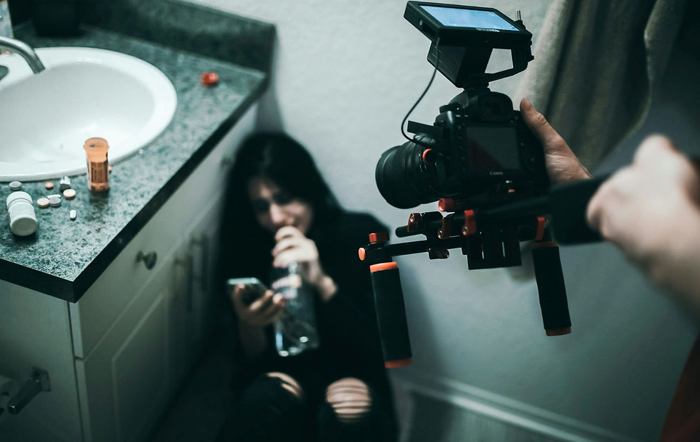 Alt text. A person sits on a bathroom floor looking distressed while holding a phone and a bottle, with various pill bottles and tablets scattered on the counter beside them. Another person films the scene using a professional camera rig, capturing the moment from above. The setting feels tense and staged for a dramatic or awareness-focused video.