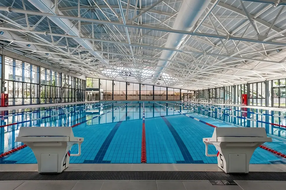 צילום אדריכלי רחב של בריכת שחייה אולימפית מקורה עם תקרת קונסטרוקציית פלדה חשופה, מקפצות זינוק בחזית וחלונות זכוכית גדולים המכניסים אור טבעי