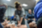 Women and teens practice CPR on mannequins in a classroom, one girl pressing chest compressions while others watch and smile.