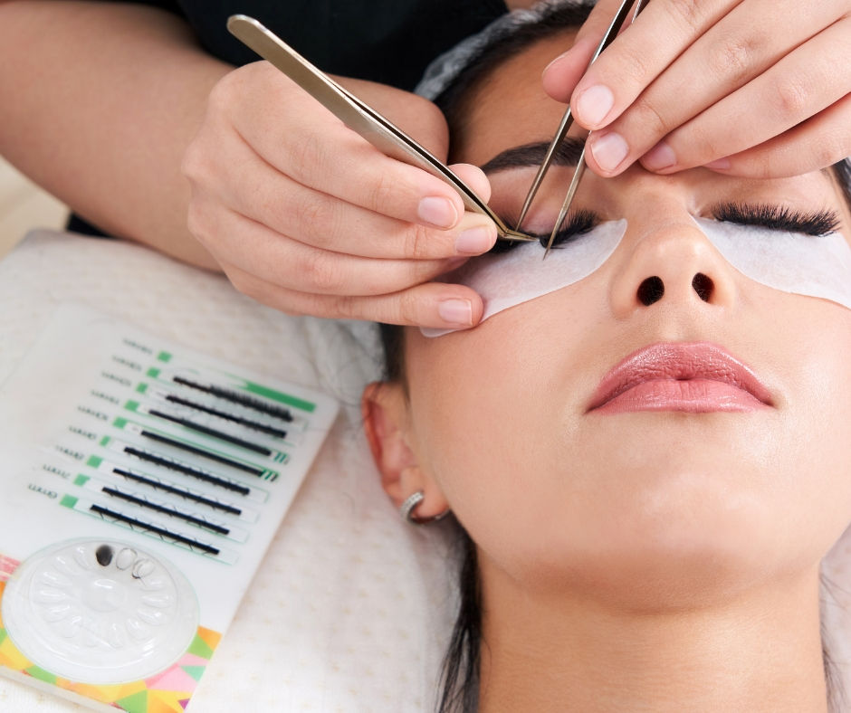 Beautician applying eyelash extensions to a woman with closed eyes, with lash trays beside her in a spa-like setting.