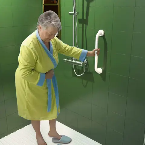 Elderly woman wearing a yellow robe holding onto a white grab bar inside a green-tiled shower, demonstrating safe entry and exit for aging-in-place bathroom design.