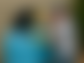 A man wearing yellow gloves and a blue cap opens the lid of a large blue residential water storage tank while cleaning it with a wooden scrub brush outside a beige house.