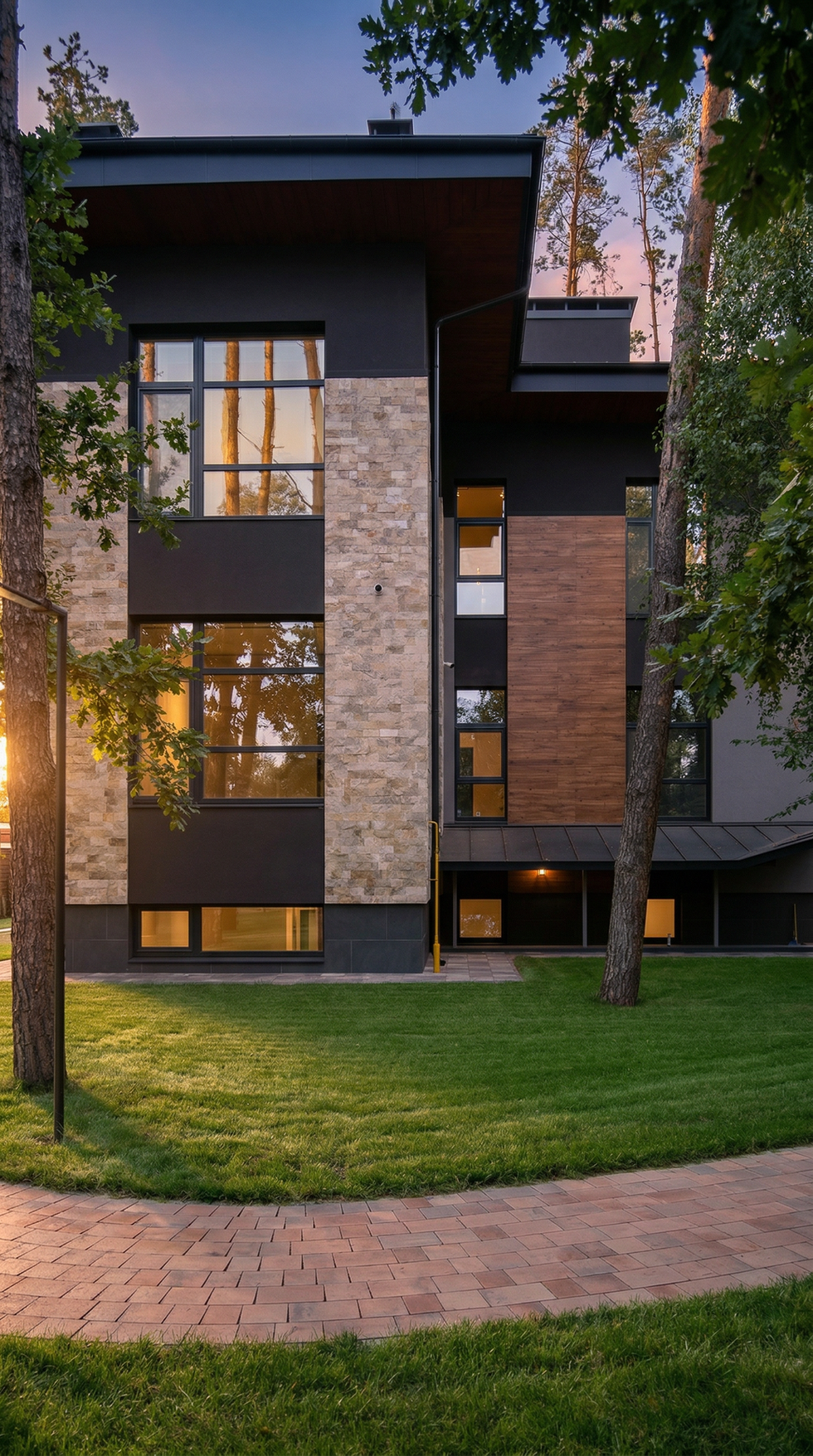 Vertical rear-side view of the mansion featuring tall windows, stone and wood materials, soft evening light, and landscaped lawn.