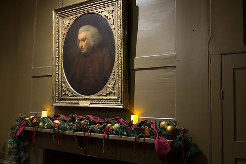 A framed portrait of Samuel Johnson hangs above a mantel decorated for Christmas with evergreen garlands, red ribbon, gold baubles, dried orange slices, and two glowing candles.