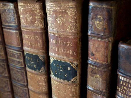 Close-up photograph of the spines of antique leather-bound books, prominently featuring two volumes of Johnson’s Dictionary with ornate gold tooling and black title labels. The aged leather shows wear and texture, evoking a sense of historical richness and scholarly tradition.