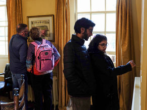 Four visitors exploring a warmly lit library room with tall windows, yellow curtains, and glass-fronted bookcases filled with old volumes.