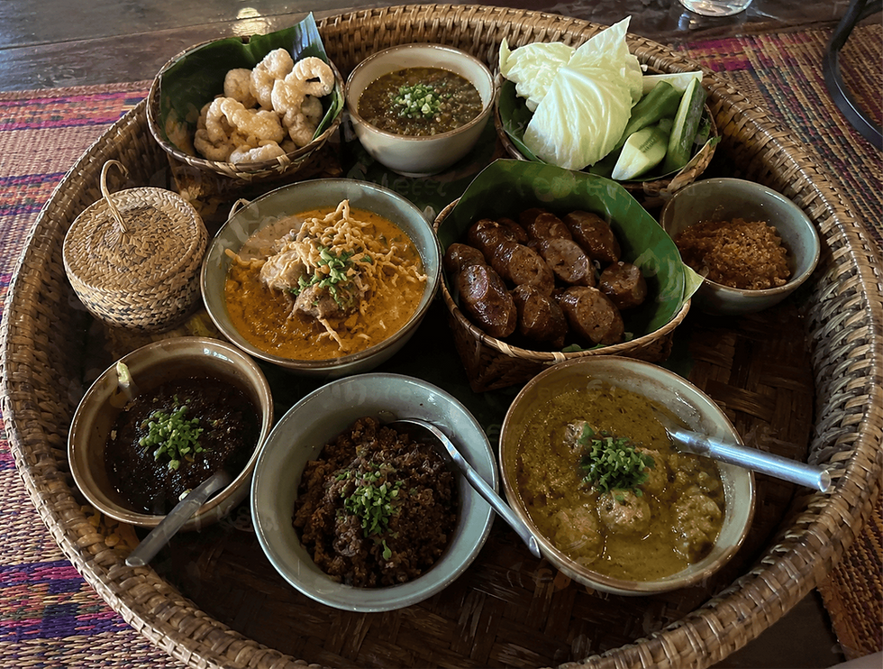 A traditional northern Thai khantoke tray with multiple dishes including khao soi, sai oua sausages, nam prik dip, pork rinds, fresh vegetables, and sticky rice in a woven basket, served on a woven rattan tray.