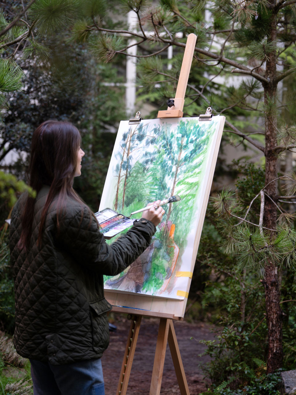 Artist stood in front of easel with large watercolour painting of trees, surrounded by forest outdoor location.