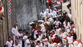 Runners wearing white and red clothing run between the high walls on Pamplona's Calle de Santo Domingo as six toros bravos and six steers charge after them.