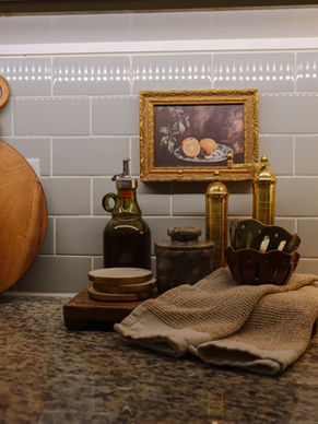 Kitchen counter with cutting boards, lemons in a bowl, oil bottle, spices, towels, painting of fruit, and lamp on a granite surface. Cozy vibe.
