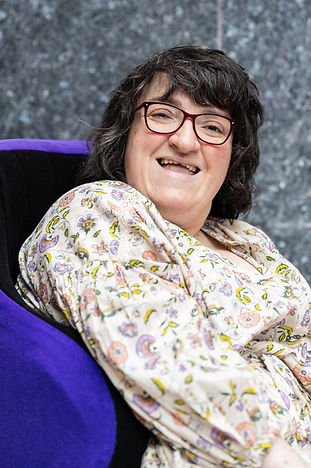 Colour photo of Kerrie. Kerrie is a white woman with short dark brown hair and red glasses. She is in a wheelchair with a purple cushion and is wearing a flowery dress. Shona Louise Photography for photo credit.