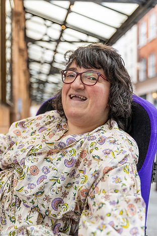 Colour photo of Kerrie. Kerrie is a white woman with short dark brown hair and red glasses. She is in a wheelchair with a purple cushion and is wearing a flowery dress. Shona Louise Photography for photo credit.