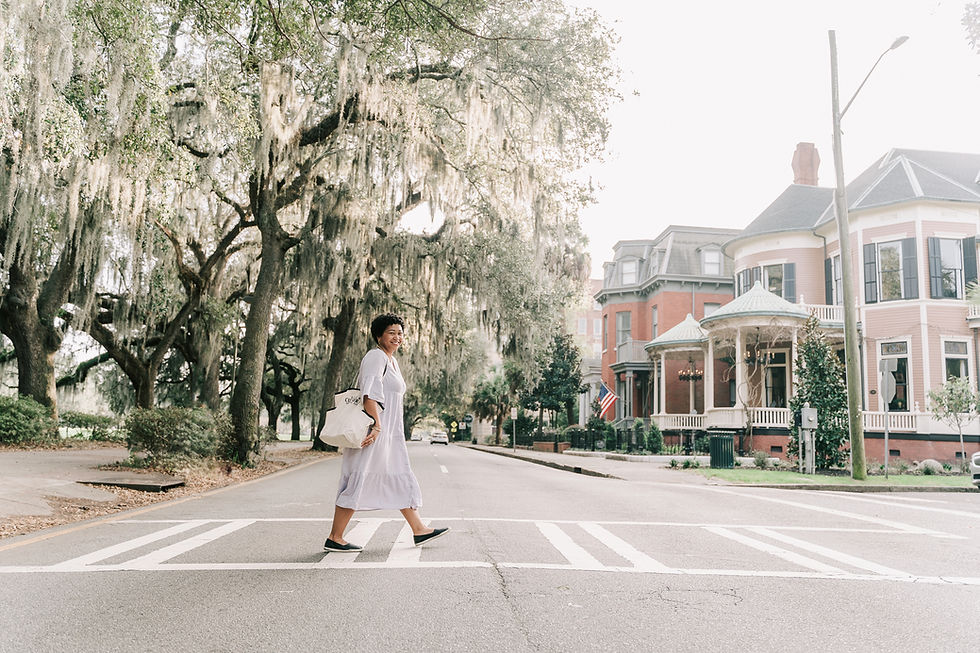 Anela strolls across a sidewalk crossing the street next to Forsyth Park in Savannah, GA with historic homes and oak trees visible in the background