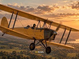 Yellow vintage biplane flies at sunset over a green valley and river, pilot visible; priAviate watermark.