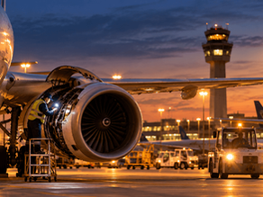 Ground crew inspects a jet engine at dusk on an airport tarmac, with a control tower and FLY logo in the background.