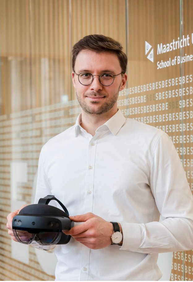 Portrait of Tim Hilken in glasses and a white shirt, holding a mixed reality headset