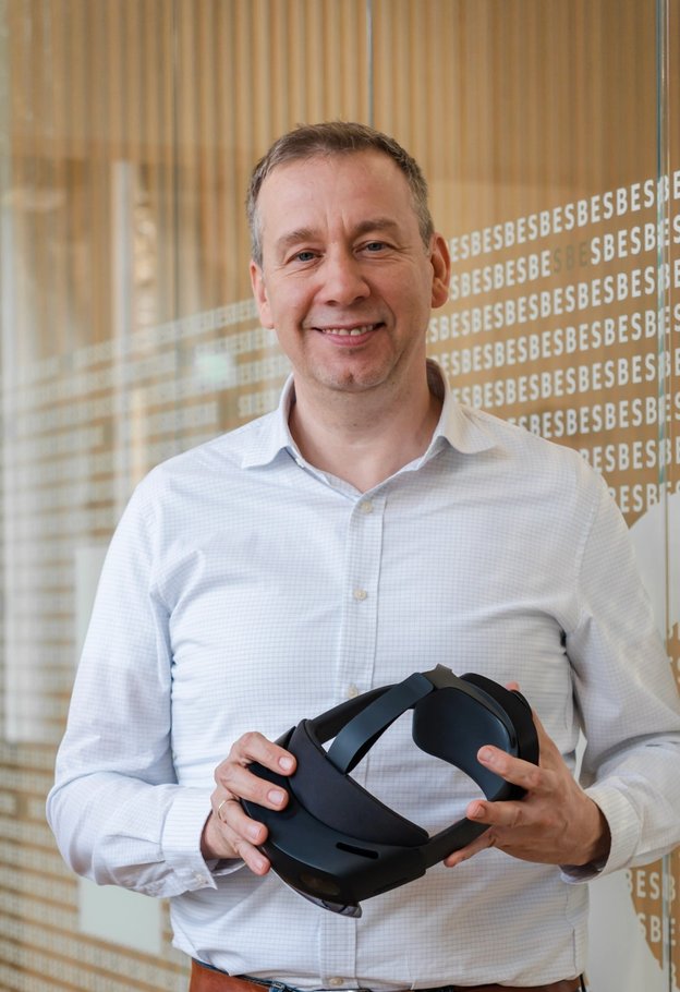 Portrait of Dominik Mahr smiling in a white shirt, holding a mixed reality headset