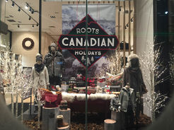 Roots holiday themed retail window display. The display shows a printed hanging banner of a snowy rocky mountain, and a hanging sign in front that reads "Roots Canadian Holidays".