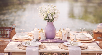 A cozy outdoor table setup with soft lavender accents, candles, and flowers beside a peaceful lakeside setting.